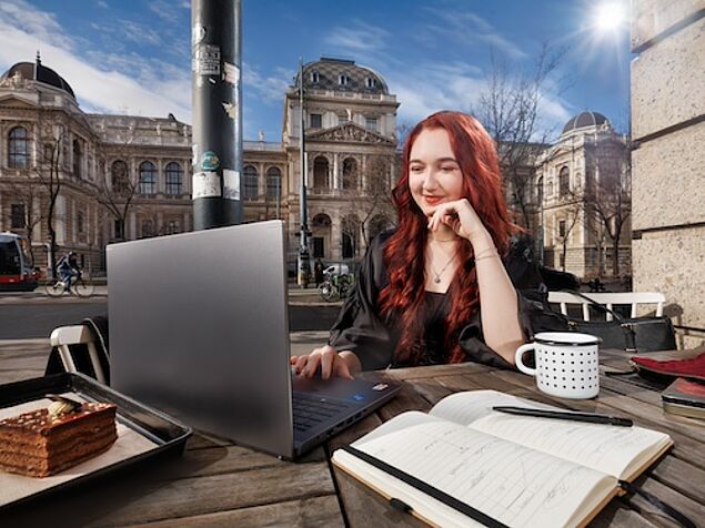 Studentin mit Lernunterlgen sitzt an einem Kaffeetisch, im Hintergrund das Hauptgebäude der Uni Wien